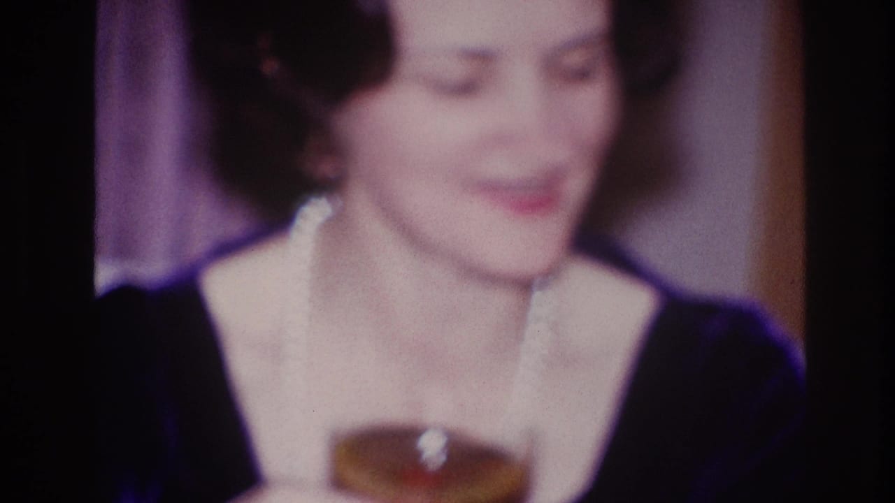 Older woman in a dark dress with a white collar smiles indoors beside a drink glass.