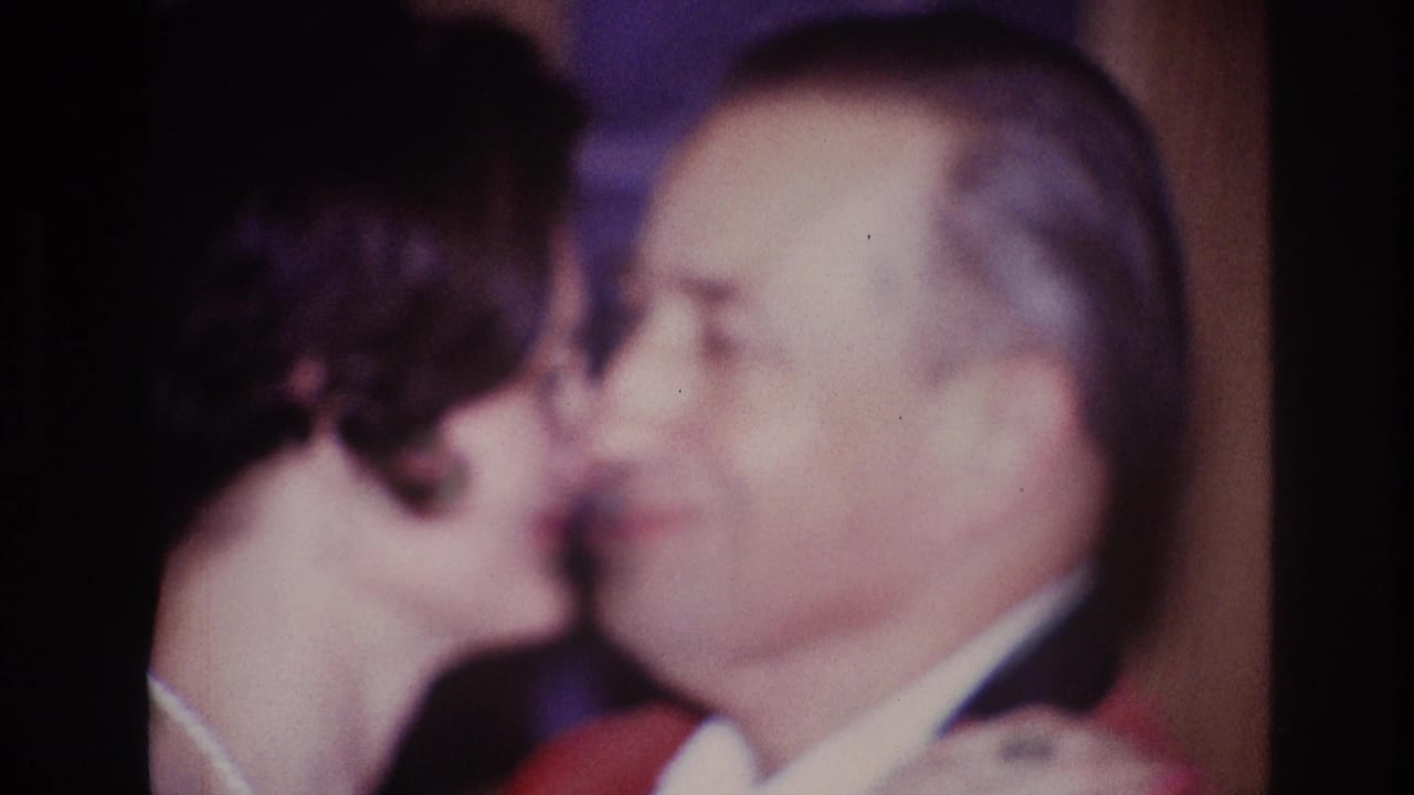 Older woman leans close to kiss an older man in a suit indoors.