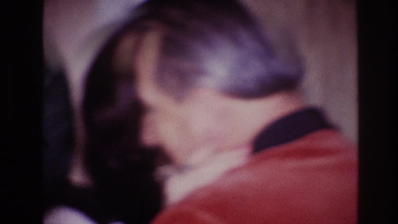Man in a red jacket embraces a woman in a dim, close-up frame.