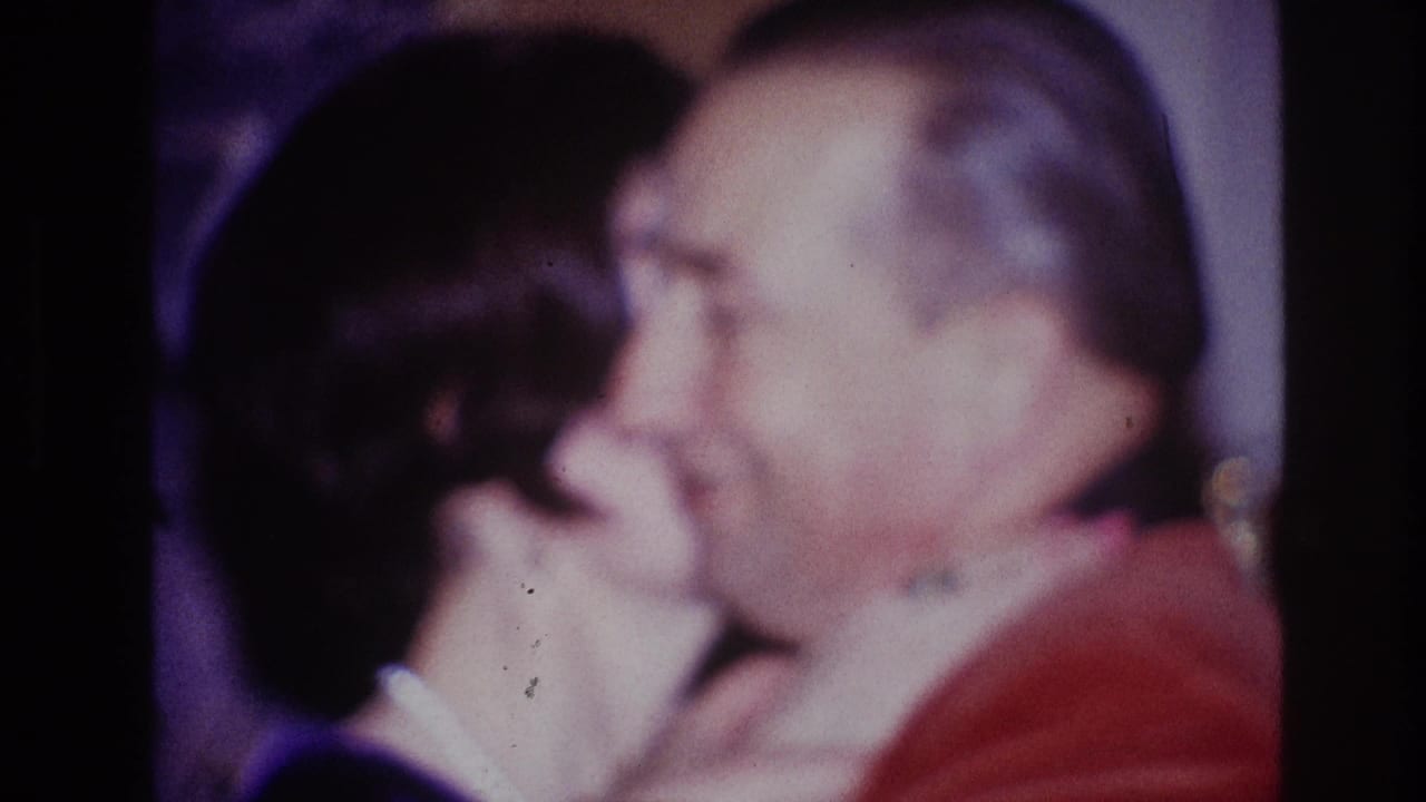 Older man in a red jacket leans toward a woman in a close embrace.