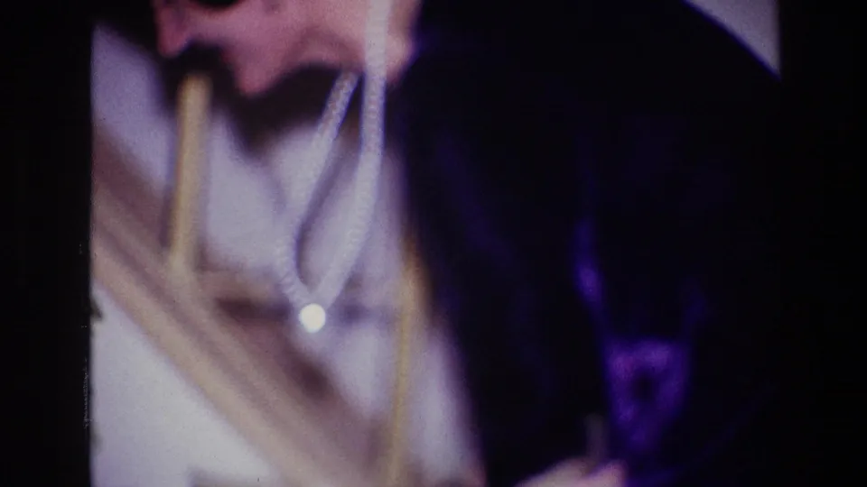 The woman in a dark dress and pearls bends forward near an indoor stair rail as the home-movie camera holds close.