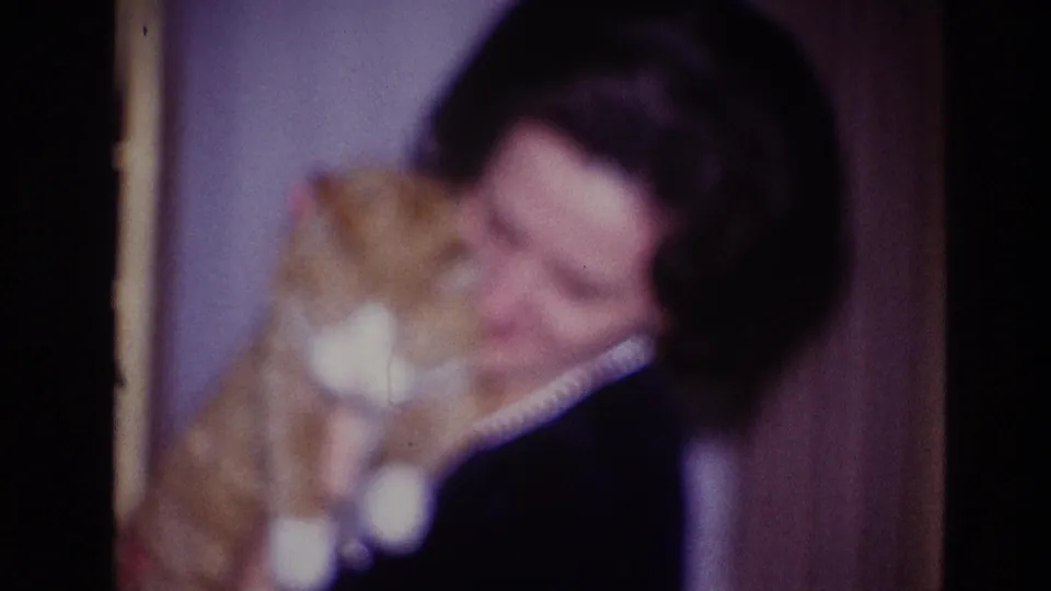 In a 1967 Ottawa home-movie frame, an adult woman wearing a pearl necklace holds an orange and white cat near her face.