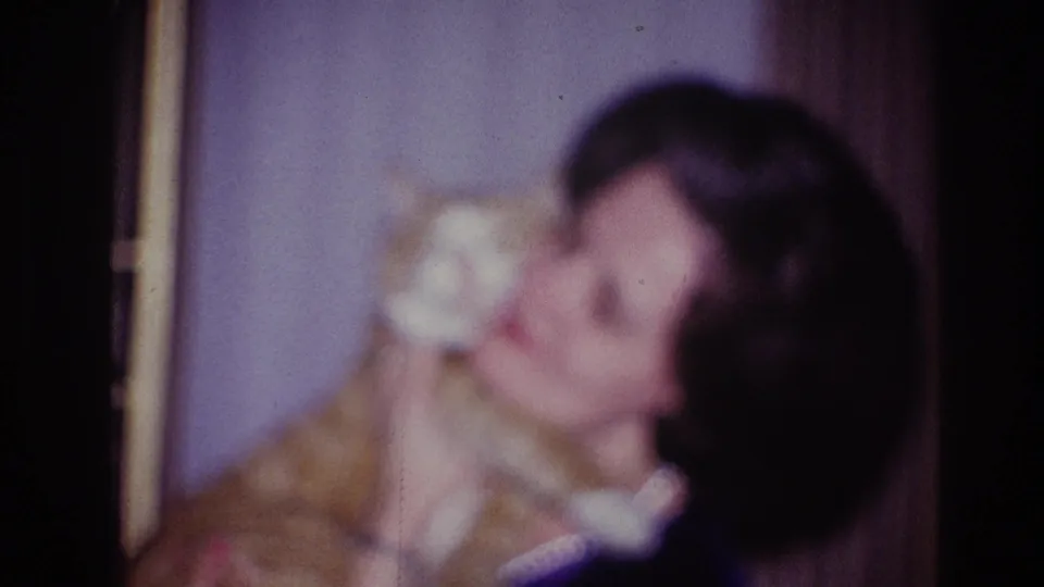 Soft-focus 8mm frame of the woman holding the orange tabby cat against her shoulder in a modest domestic room.