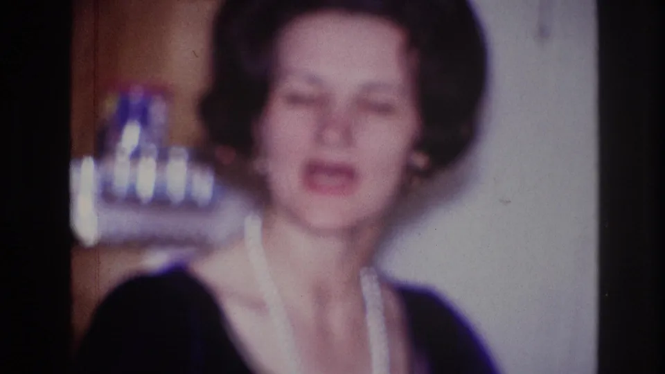 A woman with dark styled hair, red lipstick, and a pearl necklace faces the camera in a 1967 Ottawa domestic interior with shelves behind her.