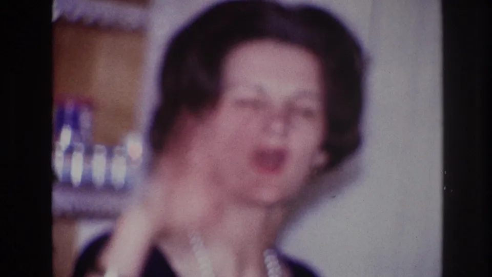 In supported 1967 Ottawa home-movie footage, an adult woman in a dark dress and pearl necklace gestures while talking indoors.
