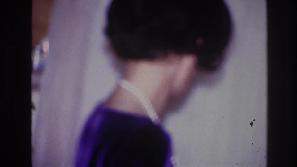 In the 1967 Ottawa home-movie setting, the woman turns away from the camera, with her pearl necklace visible against a dark dress.