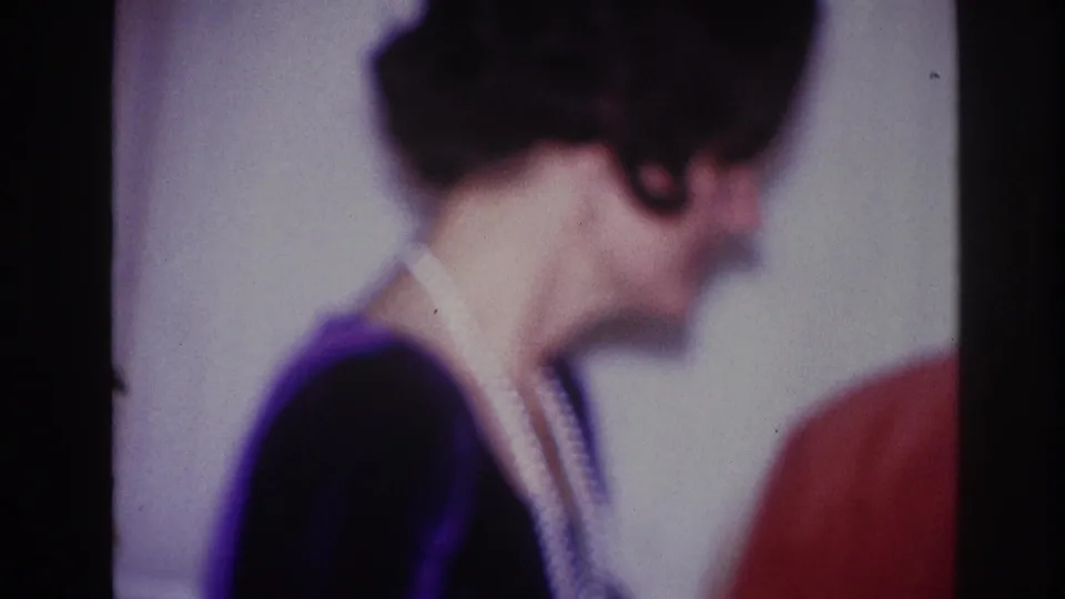 The home-movie frame shows the woman in profile, her necklace visible as she turns near another person inside the party room.