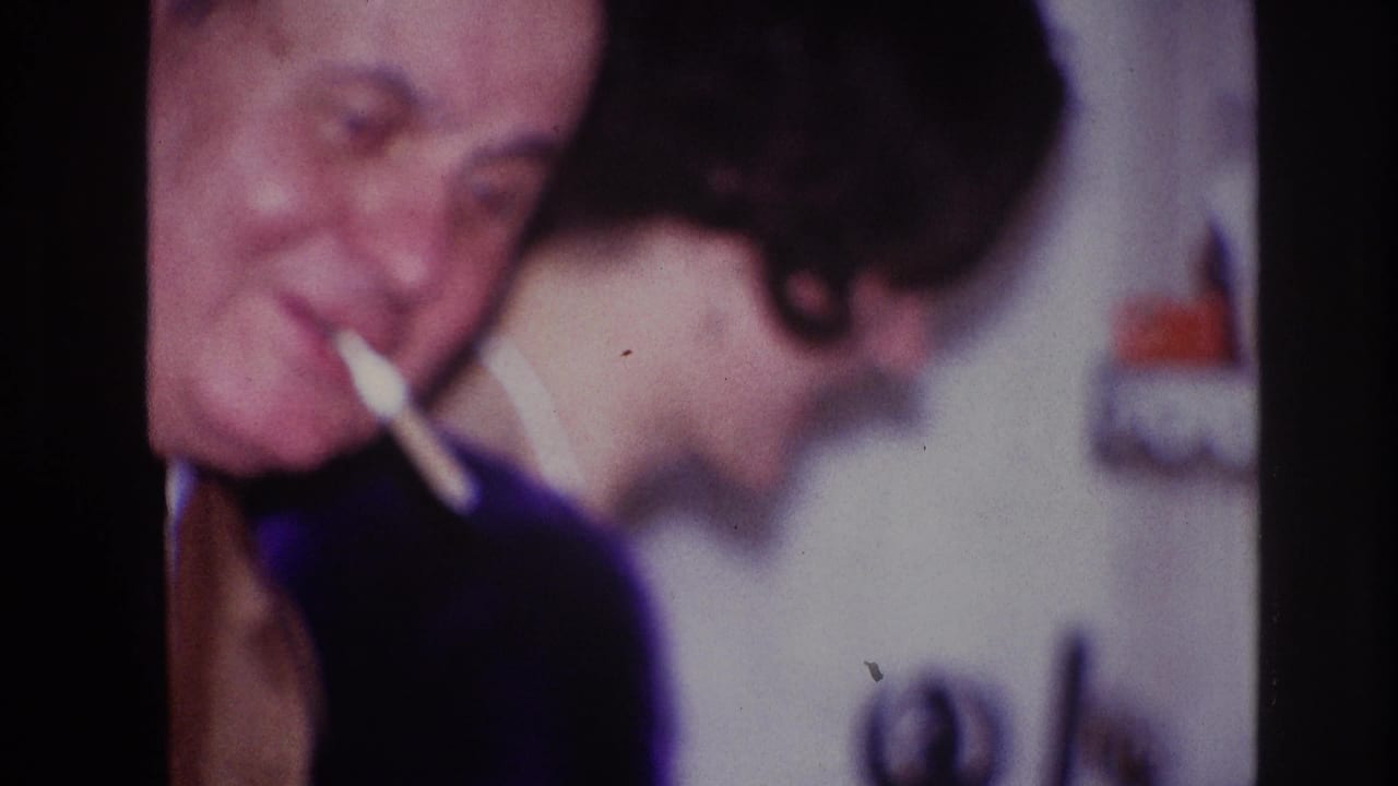 Smiling adult man with a cigarette beside a woman turned partly away indoors.