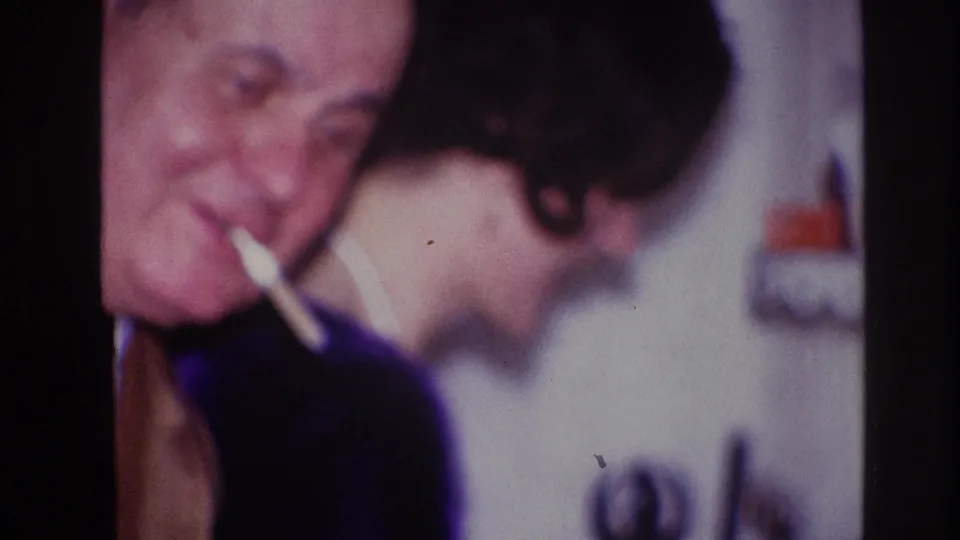1967 Ottawa home-movie frame of a smiling man holding a cigarette beside a woman in a dark top at an indoor gathering.