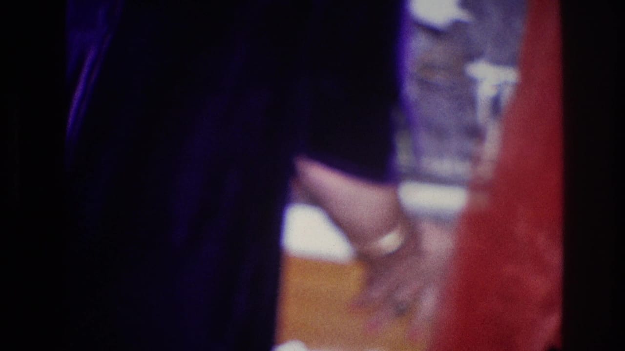 1967 Ottawa home-movie frame of a woman near a table and glass, with another figure partly visible at the edge.