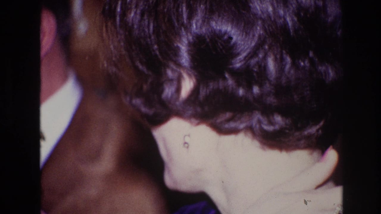 1967 Ottawa home-movie frame showing a dark-haired woman in profile with earrings near a suited man at an indoor gathering.