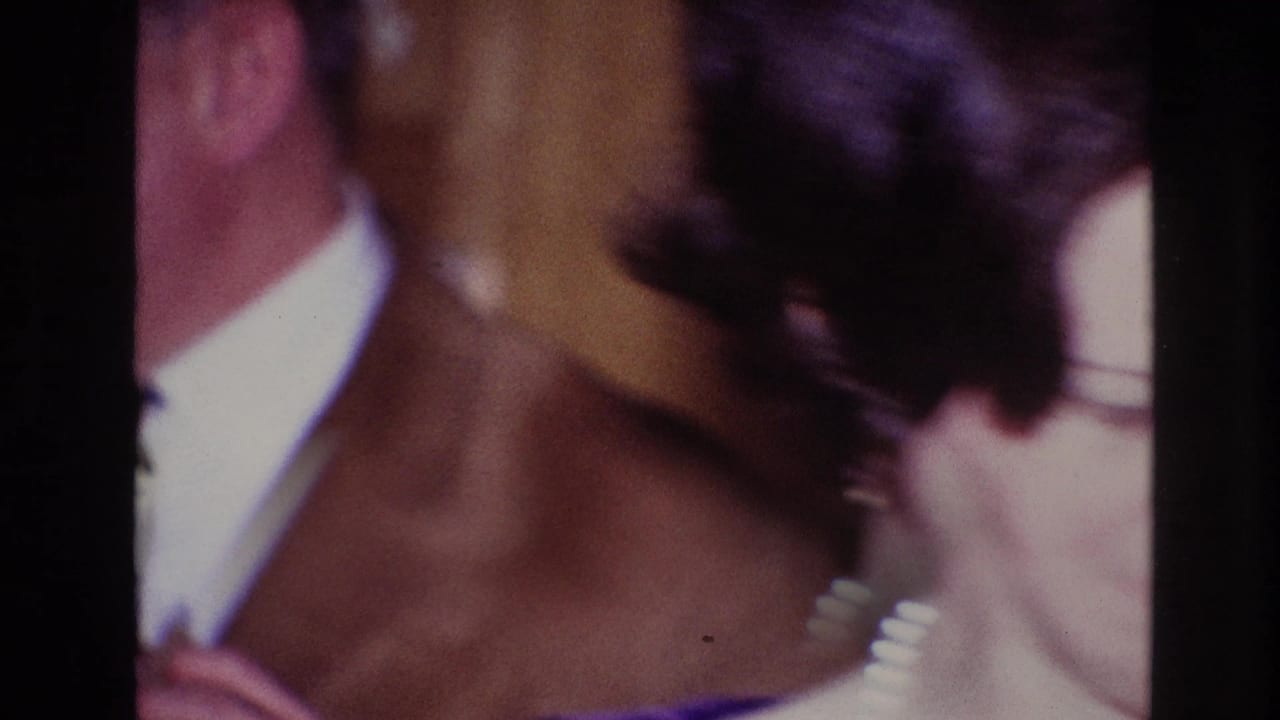 1967 Ottawa home-movie frame with a woman seen from behind wearing pearls while a suited man stands nearby indoors.