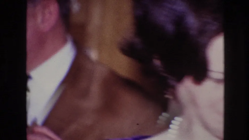 1967 Ottawa home-movie frame with a woman seen from behind wearing pearls while a suited man stands nearby indoors.