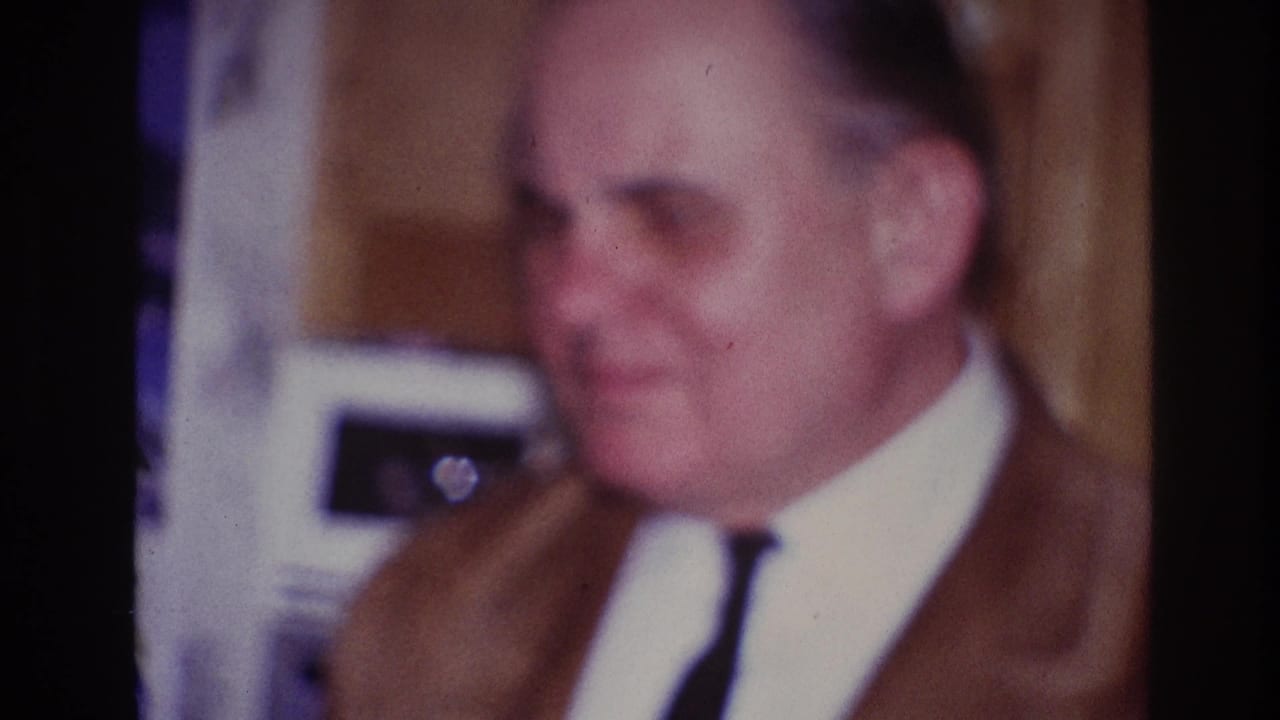 1967 Ottawa home-movie frame showing a soft-focus close-up of a man in a brown suit and tie inside a private party setting.
