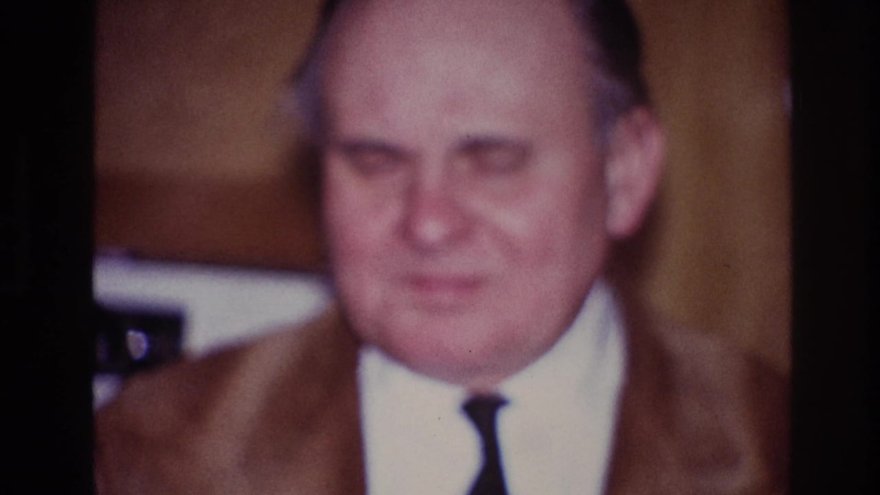 1967 Ottawa home-movie frame with a frontal close-up of a man in a brown suit and narrow tie during an indoor gathering.