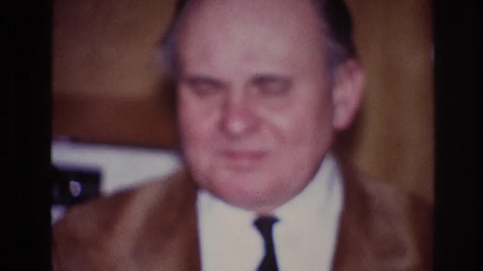 1967 Ottawa home-movie frame with a frontal close-up of a man in a brown suit and narrow tie during an indoor gathering.