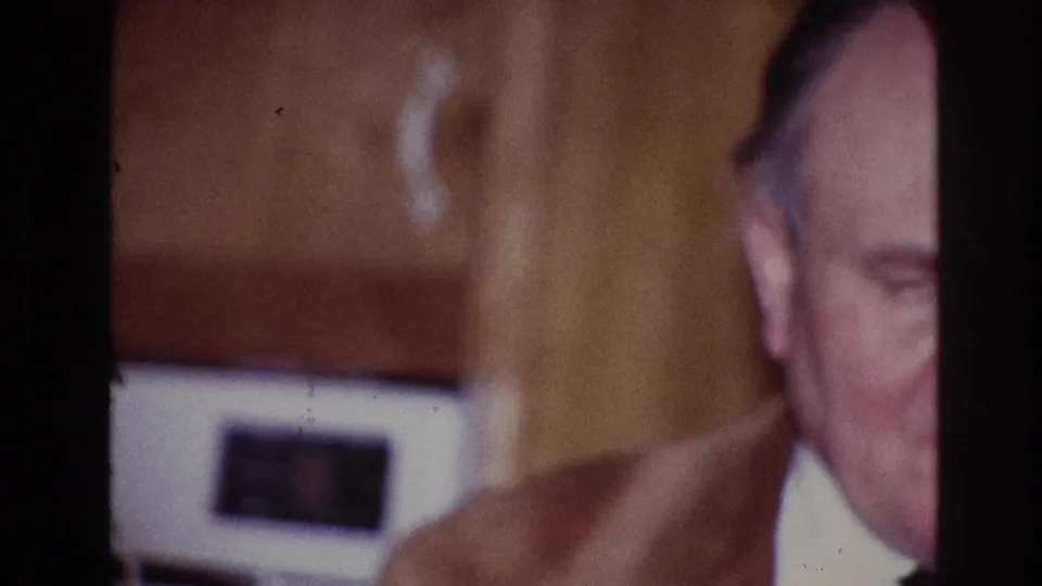 1967 Ottawa home-movie frame with an adult man partly hidden by a dark frame edge and a woman in a purple dress nearby.