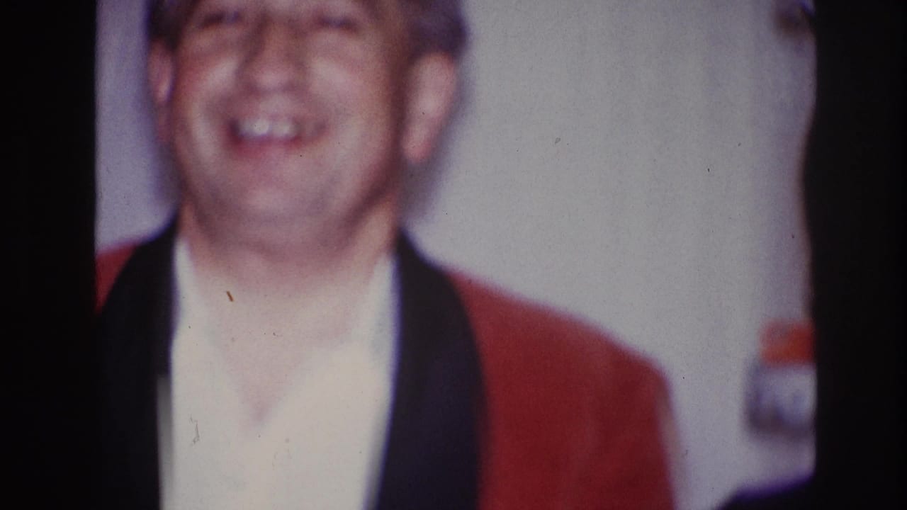 1967 Ottawa home-movie frame of an adult man in a red smoking jacket and white shirt laughing indoors.