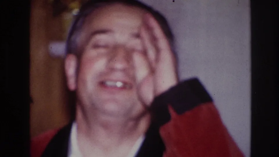 1967 Ottawa indoor frame of the man laughing with one hand over his face, wearing a red smoking jacket and white shirt.