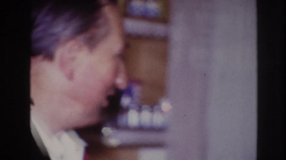 The older man turns sideways in the 1967 Ottawa interior, with shelves or objects blurred behind him.