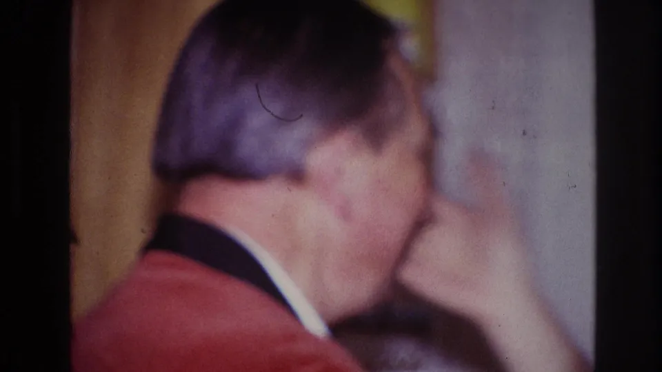The man in the red jacket appears in profile with his hand near his face during the indoor home-movie moment.