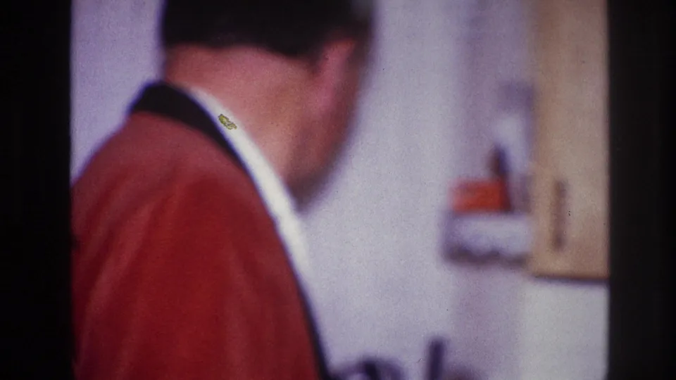 1967 Ottawa home-movie frame showing a man in a red jacket turned toward an interior wall fixture.