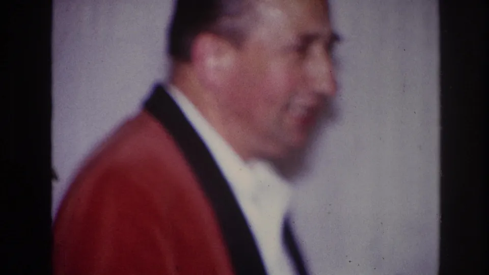 1967 Ottawa home-movie frame showing the man in soft profile, wearing a red jacket and white shirt indoors.