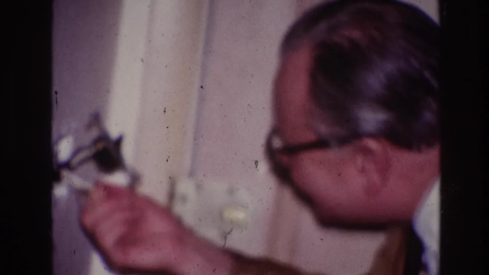 1967 Ottawa home-movie frame showing a man with glasses reaching toward a wall fixture or outlet inside a private interior.