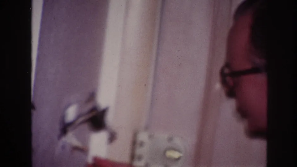 The man is partly framed in profile beside the same interior wall fixture, with the handheld 8mm image blurred.