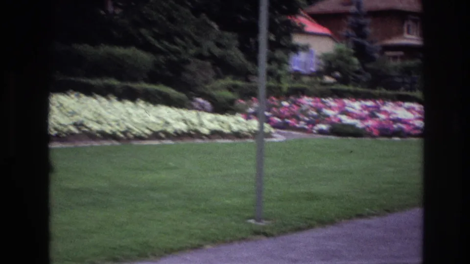 1967 Ottawa frame of a manicured lawn beside a sidewalk, with pale and pink flowers, a foreground pole, shrubs, and a partly visible house in back.