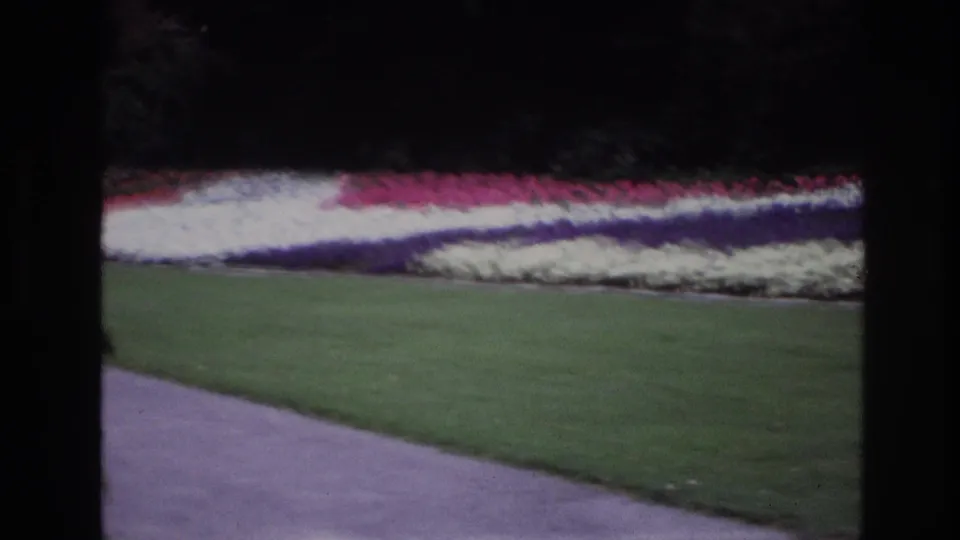 1967 Ottawa home-movie view of a paved park path, lawn, and broad red, white, and blue flower beds framed by dark film edges.