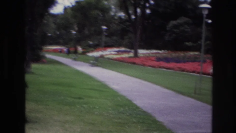 The 8mm frame shows a park path, open lawn, red and white flower beds, mature trees, lampposts, and small distant figures in Ottawa.