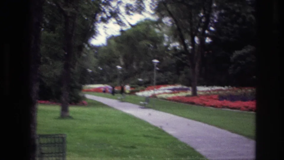 1967 Ottawa garden view with a paved path, mature trees, bright flowerbeds, and distant visitors seen through dark framing.