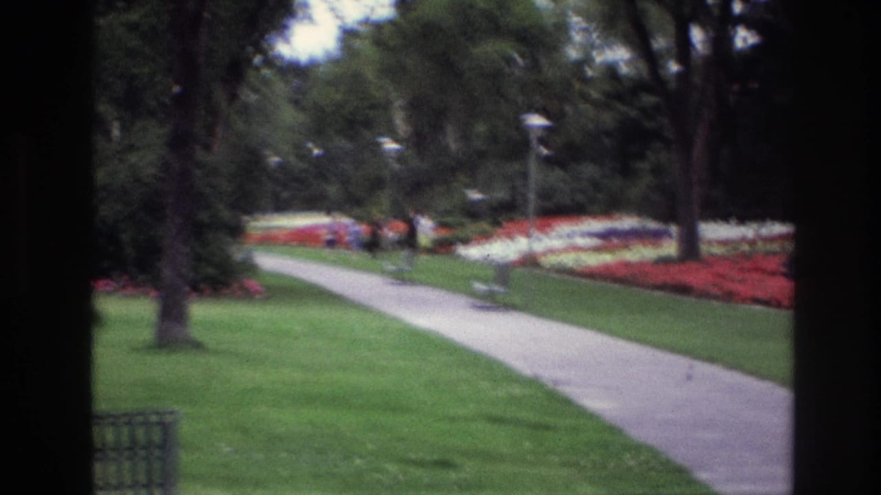Garden walkway with lawns, trees, flowerbeds, bench, and distant people.