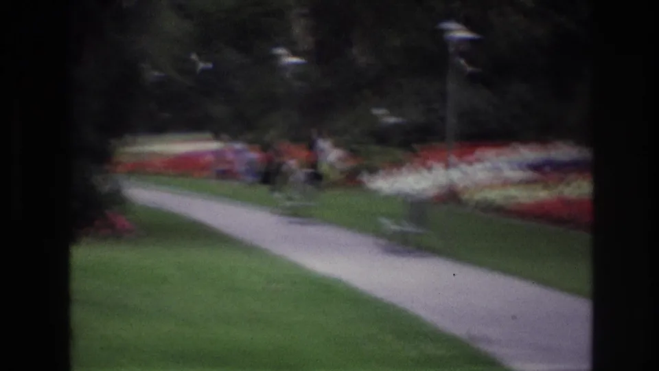 Soft-focus 1967 Ottawa garden frame showing a curving path, green lawns, red and white beds, trees, and distant visitors.