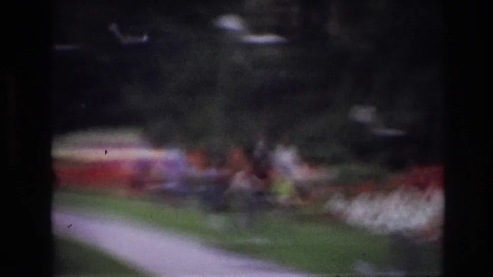 Blurry 1967 Ottawa 8mm frame showing indistinct people near a lawn, paved path, and red-and-white planting beds.
