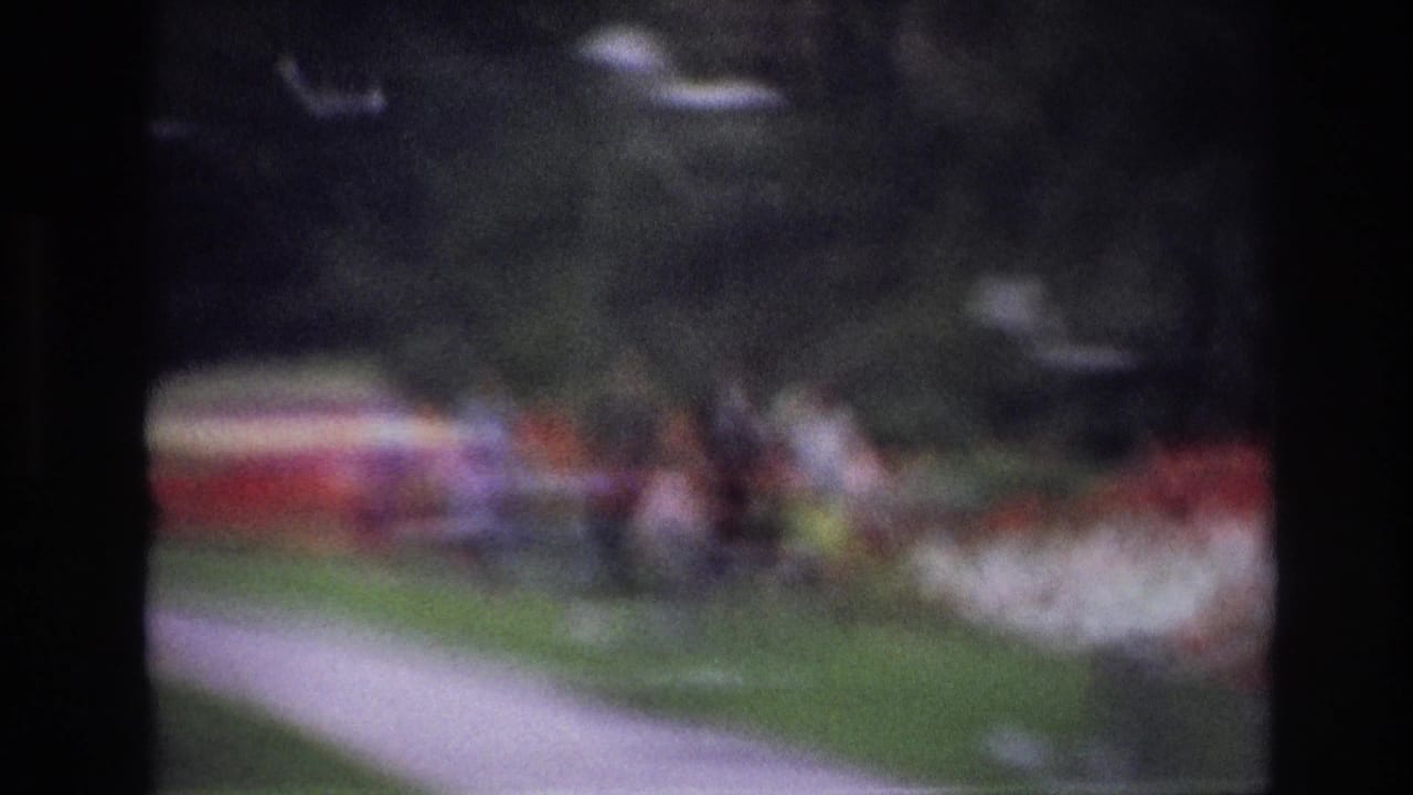 Out-of-focus 1967 Ottawa home-movie frame with soft figures beside grass, walkway, and bright garden plantings.