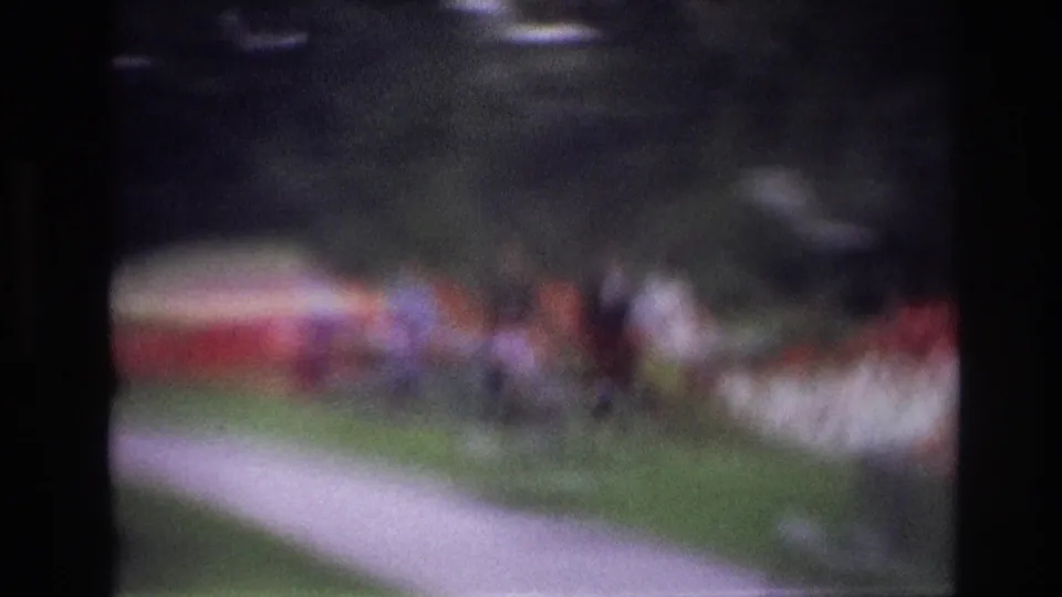Blurry Ottawa 1967 8mm frame showing a pale walkway crossing grass, with indistinct people and flower beds beyond.