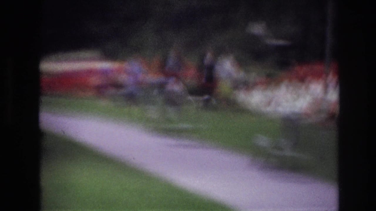 Soft 1967 Ottawa home-movie frame showing a paved park path through green grass, trees, flower beds, and indistinct people in the distance.