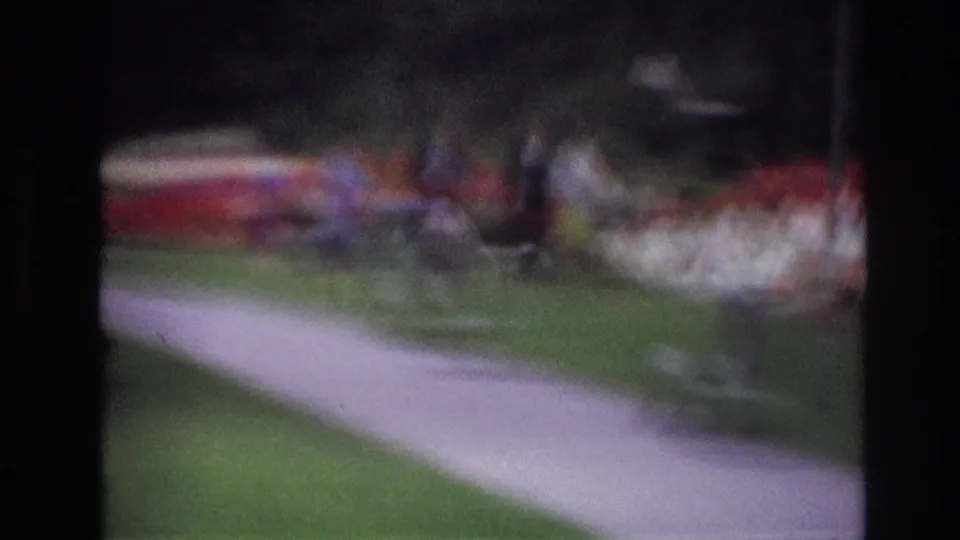 Soft 1967 Ottawa home-movie frame showing a paved park path through green grass, trees, flower beds, and indistinct people in the distance.