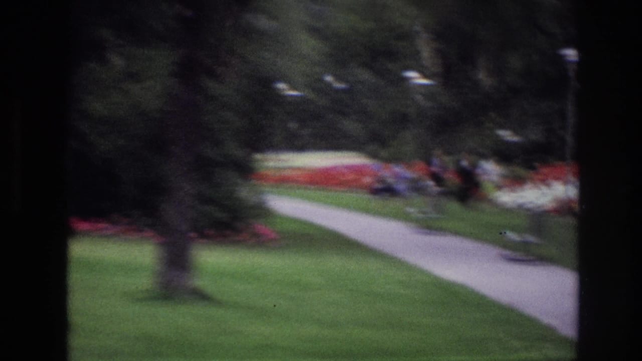 1967 Ottawa park view with a curving paved path, green lawn, trees, red and white flower beds, benches, and small distant pedestrians.