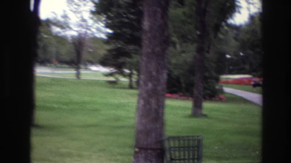 1967 Ottawa home-movie frame of a green park lawn with tree trunks, red flower beds, a path, and a dark metal waste bin in the foreground.