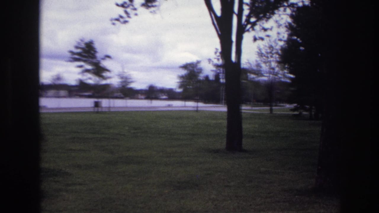 1967 Ottawa park view through a dark foreground frame, showing grass, trees, water, and distant shore under cloudy sky.