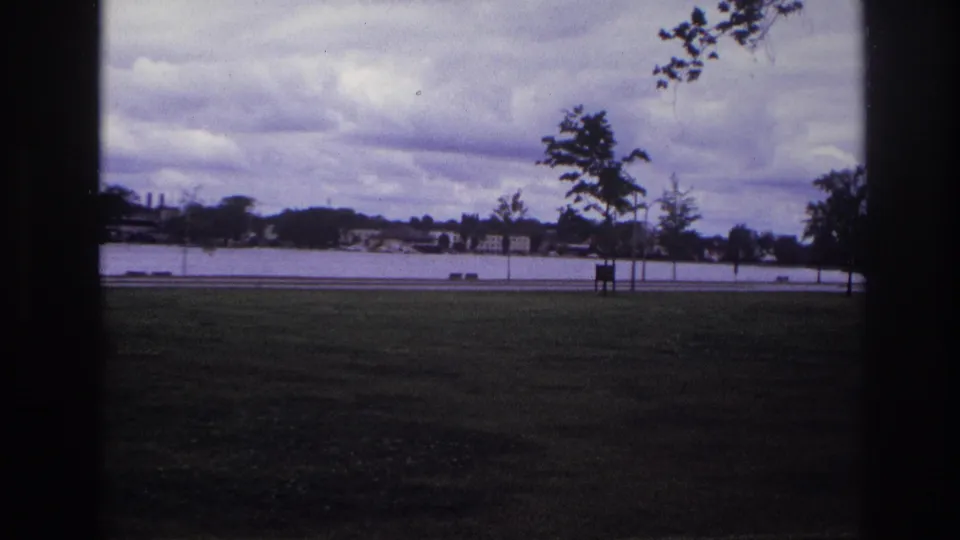 1967 Ottawa park and waterfront view showing grass, small trees, roadside objects, water, distant houses, and low clouds.