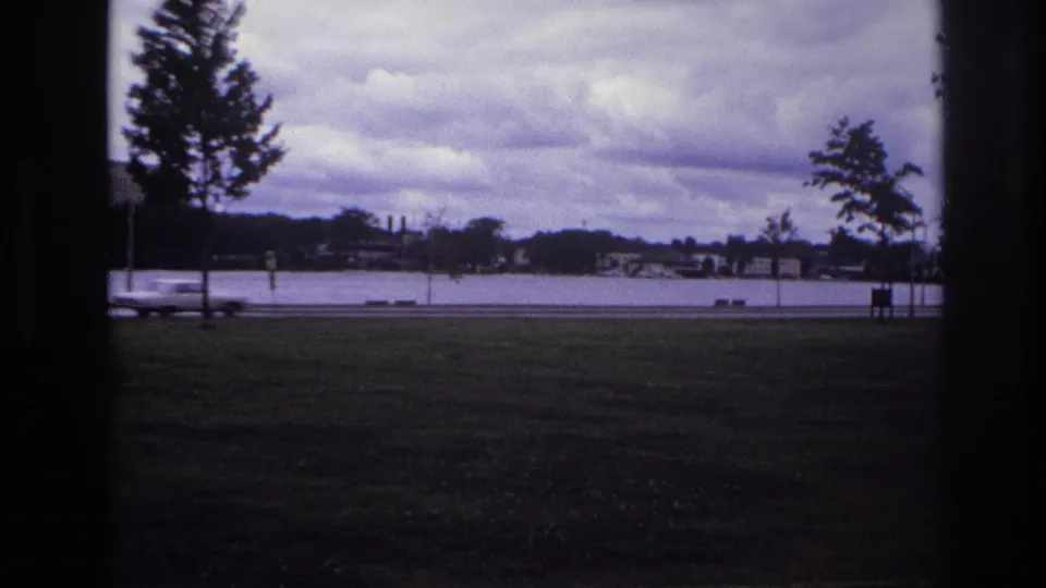Ottawa park view with a blurred car on the shoreline road, water and houses beyond, and trees under a cloudy sky.