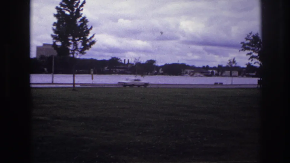 1967 Ottawa home-movie frame showing a grassy waterfront view with a young tree and a white car passing on the road.