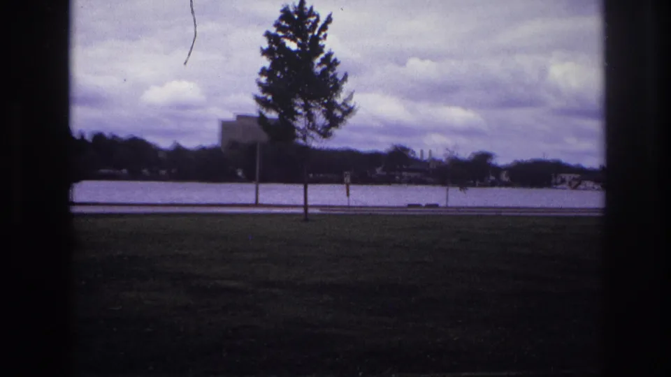 1967 Ottawa home-movie view across grass and a road toward water, trees, and low distant buildings.