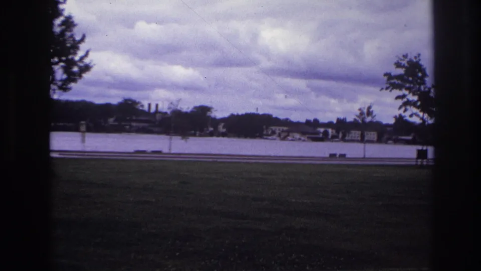 1967 Ottawa frame looking from a grassy area across a road and water toward a low distant shoreline under clouds.