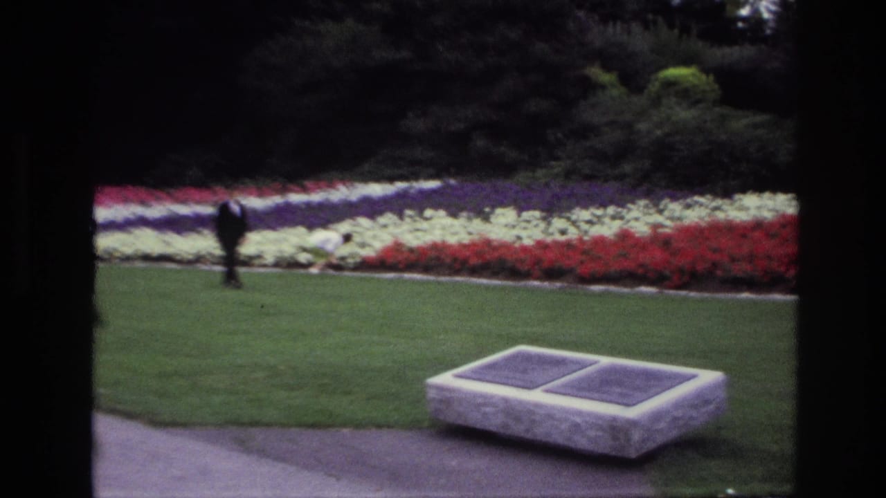 1967 Ottawa 8mm frame showing a stone memorial bench with plaques on a manicured lawn beside colorful flowerbeds and a distant visitor.