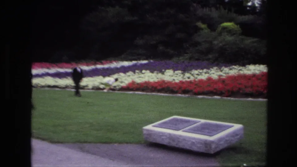 1967 Ottawa 8mm frame showing a stone memorial bench with plaques on a manicured lawn beside colorful flowerbeds and a distant visitor.